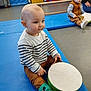 toddler, child, drum, striped_sweater, green_socks, indoor, playroom, gym_mat, floor, toy, musical_instrument, brown_pants, stacked_mats, yellow_door, curious, seated, play, baby, striped_clothing, person