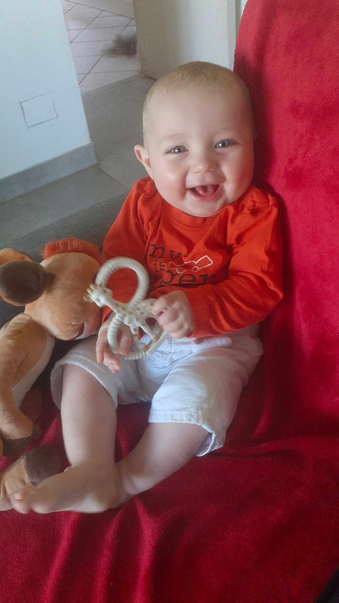 Lenny participe au concours pour gagner de l'argent avec cette photo : baby, child, couch, cute, face, foot, giraffe_teether, hand, happy, home, indoor, infant, orange_shirt, person, plush_toy, red_blanket, sitting, smiling, toy, white_shorts