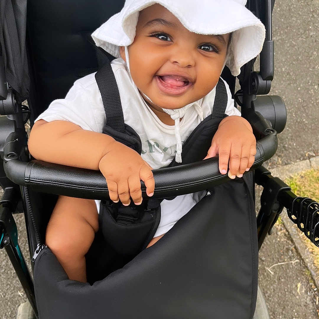 Guled is registered to the contest to win money with this photo: baby, baseballcap, bonnet, cap, clothing, coat, face, happy, hat, head, hood, jacket, machine, person, photography, portrait, smile, stroller, sunhat, wheel