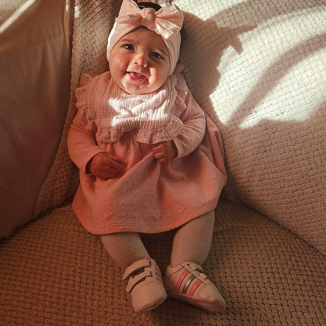 Inaya a rejoint le concours — aidez-le/la à gagner de superbes lots ! baby, child, clothing, couch, cozy, footwear, happy, headband, home, indoor, infant, person, pink_dress, seated, shadow, smiling, soft_lighting, sunlight, texture, warm_colors