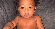 Jaxxon is registered to the contest to win money with this photo: child, toddler, curly_hair, indoor, couch, gray_furniture, bare_chest, baby_skin, curious_expression, large_eyes, hand, seated, living_room, furniture, portrait, young_child, home, person, infant, casual