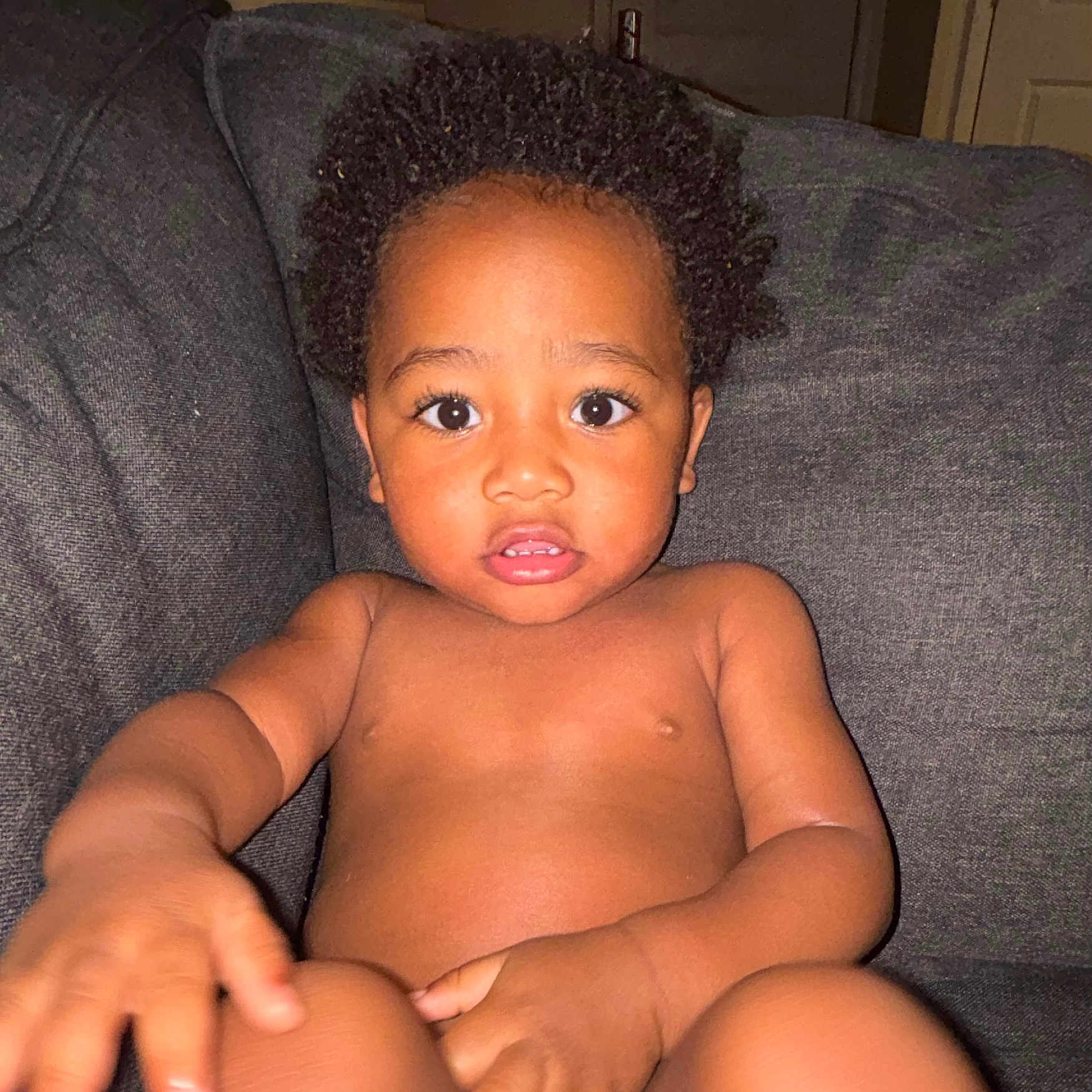 Jaxxon is registered to the contest to win money with this photo: child, toddler, curly_hair, indoor, couch, gray_furniture, bare_chest, baby_skin, curious_expression, large_eyes, hand, seated, living_room, furniture, portrait, young_child, home, person, infant, casual