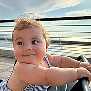 Charli participe au concours pour gagner de l'argent avec cette photo : arm, baby, blue_eyes, child, closeup, clouds, curly_hair, daylight, face, happy, metal, outdoor, person, railing, seat, sky, smiling, striped_clothing, sunlight, waterfront