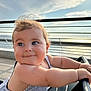 arm, baby, blue_eyes, child, closeup, clouds, curly_hair, daylight, face, happy, metal, outdoor, person, railing, seat, sky, smiling, striped_clothing, sunlight, waterfront