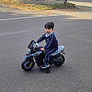 Tim Yousif joined the competition — help win amazing prizes! child, toy_motorcycle, tracksuit, black_shoes, asphalt, suburban, trees, bushes, house, curious, outdoor, daylight, road, training_wheels, person, young_child, play, vehicle, sidewalk, residential