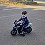 child, toy_motorcycle, tracksuit, black_shoes, asphalt, suburban, trees, bushes, house, curious, outdoor, daylight, road, training_wheels, person, young_child, play, vehicle, sidewalk, residential