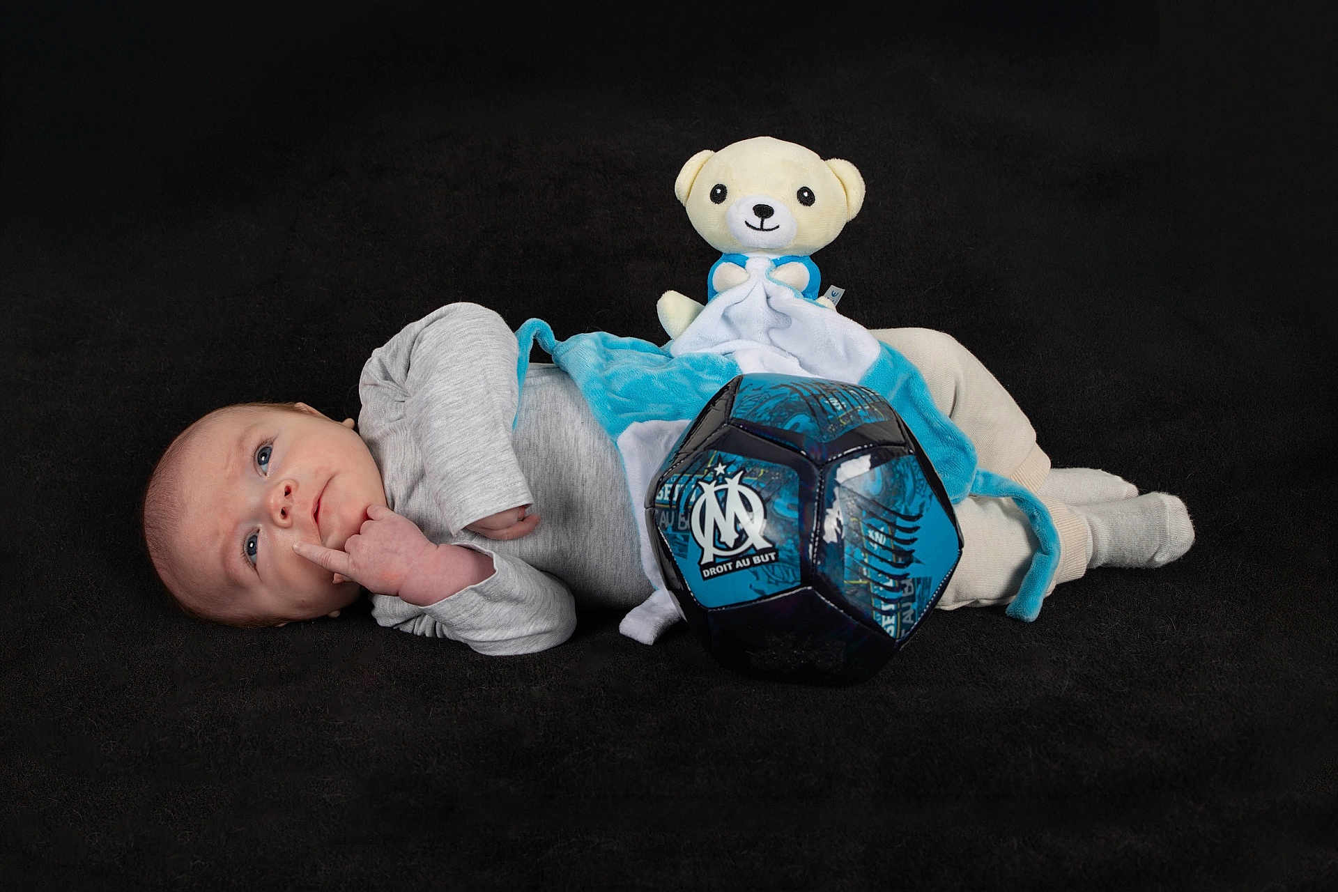 Paul participe au concours pour gagner de l'argent avec cette photo : baby, infant, newborn, teddy_bear, stuffed_toy, soccer_ball, blue_blanket, onesie, lying_down, portrait, studio_shot, black_background, curious_expression, hand_to_face, socks, soft_toy, cozy, props, blue, photography