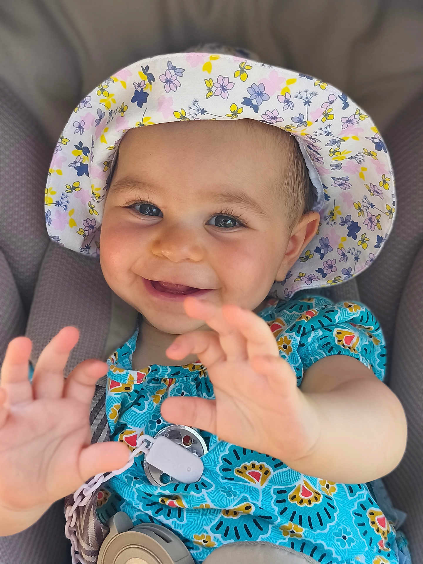 Salomé participe au concours pour gagner de l'argent avec cette photo : baby, child, smiling, hat, floral_hat, blue_dress, colorful_clothing, happy, hands, face, cute, infant, seat, car_seat, patterned_clothing, closeup, portrait, young_child, skin, person