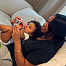 child, toddler, mother, parent, family, cuddling, sitting, lying_down, smiling, portrait, indoors, couch, pillows, bedroom, home, black_shirt, curly_hair, adult, playing_cards, cozy