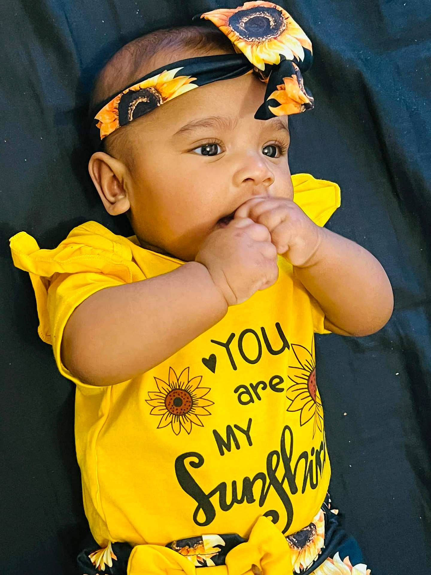 adorable, baby, baby_fashion, black_background, bright_colors, chewing, child, close_up, clothing, cute, eyes, face, hands, headband, infant, lying_down, portrait, skin, sunflower, yellow_shirt