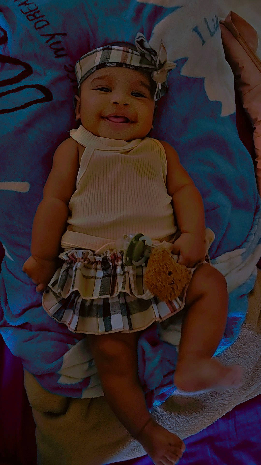 baby, blanket, child, clothing, comfort, cute, foot, hand, happy, headband, indoors, infant, lying_down, pacifier, person, plaid_skirt, relaxed, skin, smiling, toy