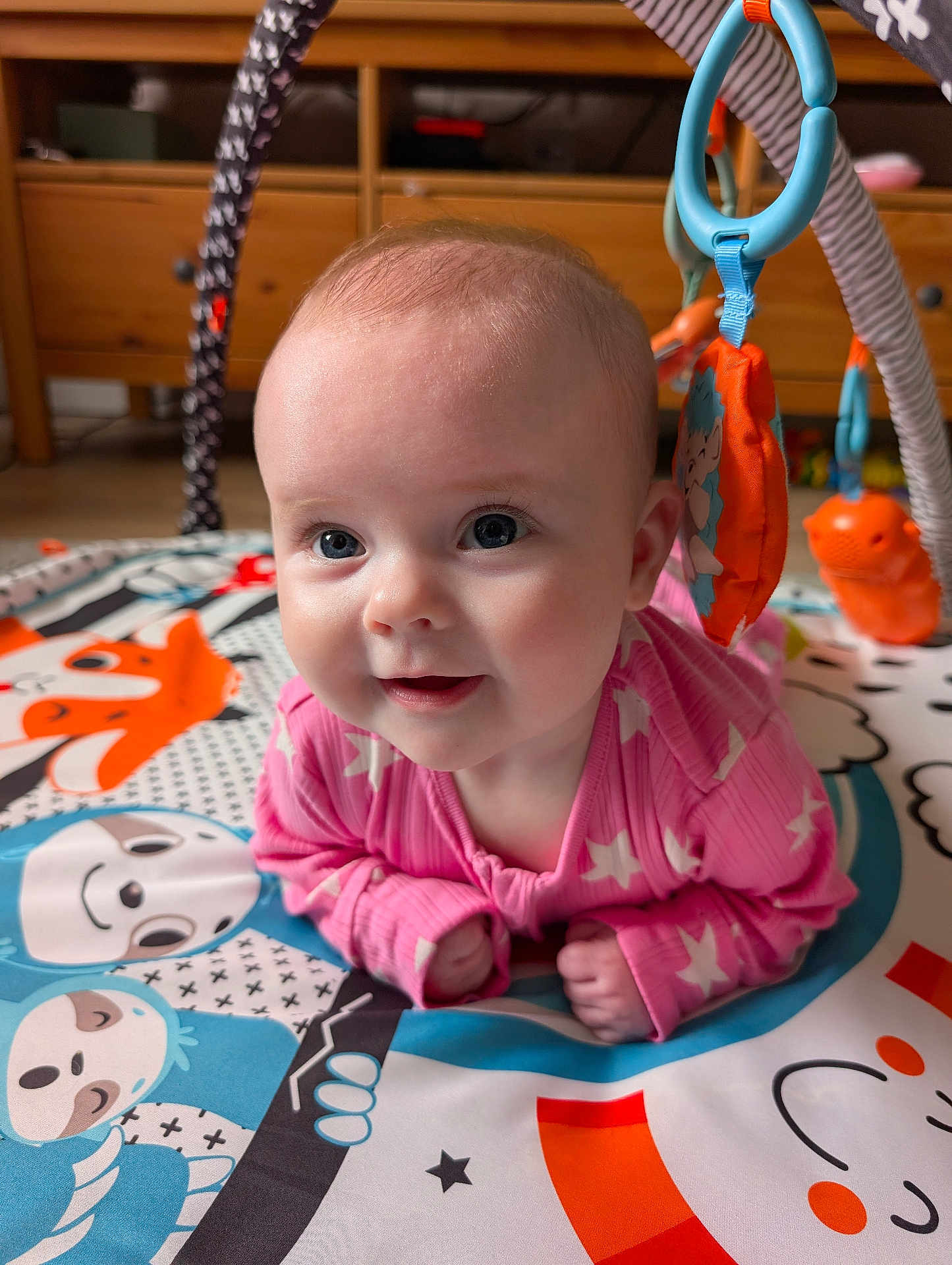 Sophia is registered to the contest to win money with this photo: baby, infant, pink_clothing, play_mat, toy, animal_design, blue, orange, cute, happy, face, child, indoor, wooden_furniture, smiling, colorful, toddler, crawling, playtime, closeup