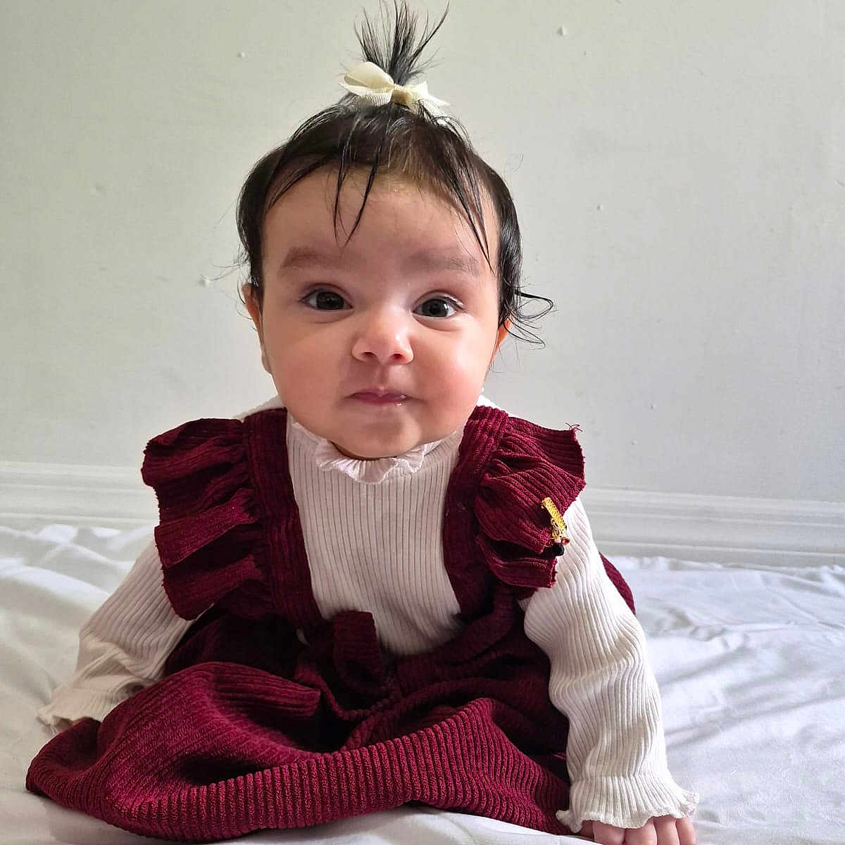 Paola is registered to the contest to win money with this photo: baby, burgundy_dress, child, curious_expression, cute, cute_outfit, face, hair_bow, indoors, infant, person, portrait, sitting, small_hands, smiling, soft_lighting, toddler, white_background, white_shirt, young_child