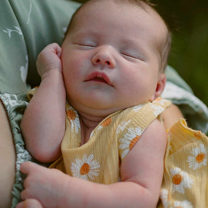 Verena is registered to the contest to win money with this photo: arm, baby, child, close_up, daisy_pattern, expression, face, hand, holding, infant, nature, outdoor, peaceful, person, portrait, relaxation, skin, sleeping, soft_focus, yellow_dress