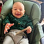 baby, smiling, green_shirt, grey_pants, high_chair, indoor, happy, child, face, hand, book, card, laundry_basket, clothing, seat, person, portrait, furniture, expression, home