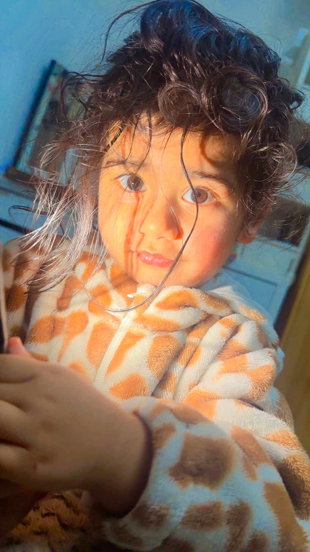 Guntas joined the competition — help win amazing prizes! child, face, hair, fleece, giraffe_pattern, jacket, indoor, warm_light, portrait, young, curly_hair, cozy, expression, hand, brown_eyes, rosy_cheeks, casual_clothing, home, blurry_background, curiosity