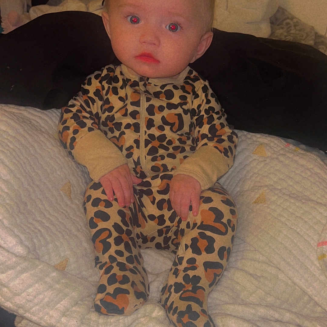 Xiomara joined the competition — help win amazing prizes! baby, bed, blanket, child, clothing, cushion, cute, expression, face, hair_clip, indoor, infant, leopard_print, onesie, person, portrait, puma_bag, red_eye, sitting, soft_lighting