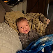 Michael is registered to the contest to win money with this photo: baby, blanket, blue_blanket, brown_blanket, child, clothing, comfort, couch, cozy, face, happy, home, indoors, infant, portrait, quilt, relaxing, smiling, soft_lighting, warm