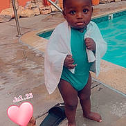 Kareem is registered to the contest to win money with this photo: child, toddler, pool, swimming_pool, towel, swimsuit, outdoor, portrait, standing, face, eyes, curly_hair, dark_skin, baby, water, concrete, potted_plant, rocks, sandal, heart_sticker