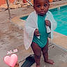 child, toddler, pool, swimming_pool, poolside, towel, swimsuit, wet, outdoor, portrait, standing, looking_at_camera, serious_expression, curly_hair, dark_skin, barefoot, flip_flop, adult_foot, plants, rocks