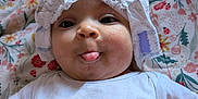 Naomi joined the competition — help win amazing prizes! baby, child, face, tongue, white_clothing, headwear, blanket, floral_pattern, playful, cheeks, lying_down, cute, infant, indoors, pacifier_clip, pink, expression, person, closeup, soft_lighting