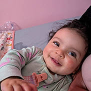 Naomi joined the competition — help win amazing prizes! baby, child, smile, pajamas, hand, face, bed, blanket, pink_wall, cute, infant, happy, indoors, person, soft_lighting, closeup, portrait, cozy, expression, lying_down