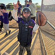 Mason is registered to the contest to win money with this photo: child, baseball, catcher, helmet, glove, protective_gear, sports, field, fence, sunlight, shadow, team, uniform, outdoor, sport_equipment, young_player, kids_sports, recreation, fun, game