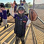 child, baseball, catcher, helmet, glove, protective_gear, sports, field, fence, sunlight, shadow, team, uniform, outdoor, sport_equipment, young_player, kids_sports, recreation, fun, game
