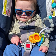 Matthew is registered to the contest to win money with this photo: baby, child, sunglasses, car_seat, toy, blue_pants, jacket, sunlight, straps, seatbelt, infant, cute, hands, face, expression, indoors, safety, plastic_toys, closeup, portrait