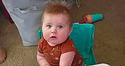 Lorrin joined the competition — help win amazing prizes! baby, high_chair, carpet, toy, laundry_basket, furniture, indoor, child, curious, person, clothing, seat, hair, face, hand, floor, living_room, blue_eyes, orange_shirt, expression