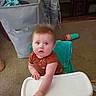 baby, high_chair, carpet, toy, laundry_basket, furniture, indoor, child, curious, person, clothing, seat, hair, face, hand, floor, living_room, blue_eyes, orange_shirt, expression