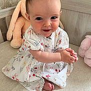 Anna is registered to the contest to win money with this photo: baby, child, smiling, dress, clapping, plush_toy, bed, cute, happy, indoor, person, toddler, patterned_clothing, foot, hand, face, head, furniture, soft_toy, infant