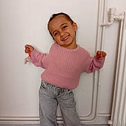 Samira a rejoint le concours — aidez-le/la à gagner de superbes lots ! casual_clothing, child, cute, face, floor, hair, happy, head, indoor, jeans, person, pink_sweater, playful, pose, radiator, smiling, standing, toy, wall, young_child