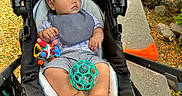 Maverick is registered to the contest to win money with this photo: baby, child, stroller, outdoor, park, pathway, tree, leaves, toys, ball, sitting, portrait, face, serious, infant, clothing, bib, striped_shorts, bare_feet, sunlight
