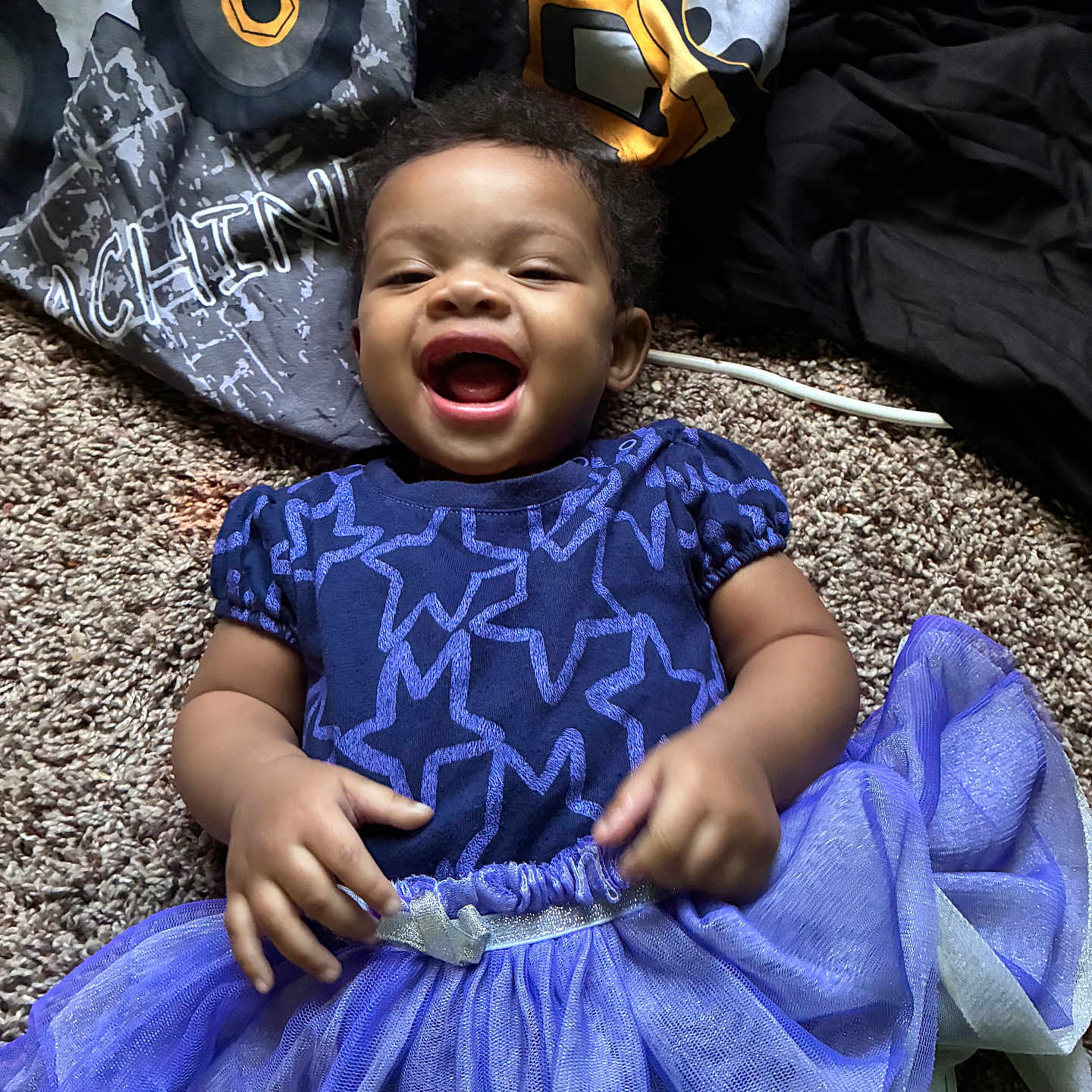 Khloe Grace is registered to the contest to win money with this photo: baby, child, smiling, laughing, blue_dress, tutu_skirt, carpet, blanket, fabric, happy, infant, cute, playful, clothing, person, portrait, indoor, young_child, star_pattern, toddler
