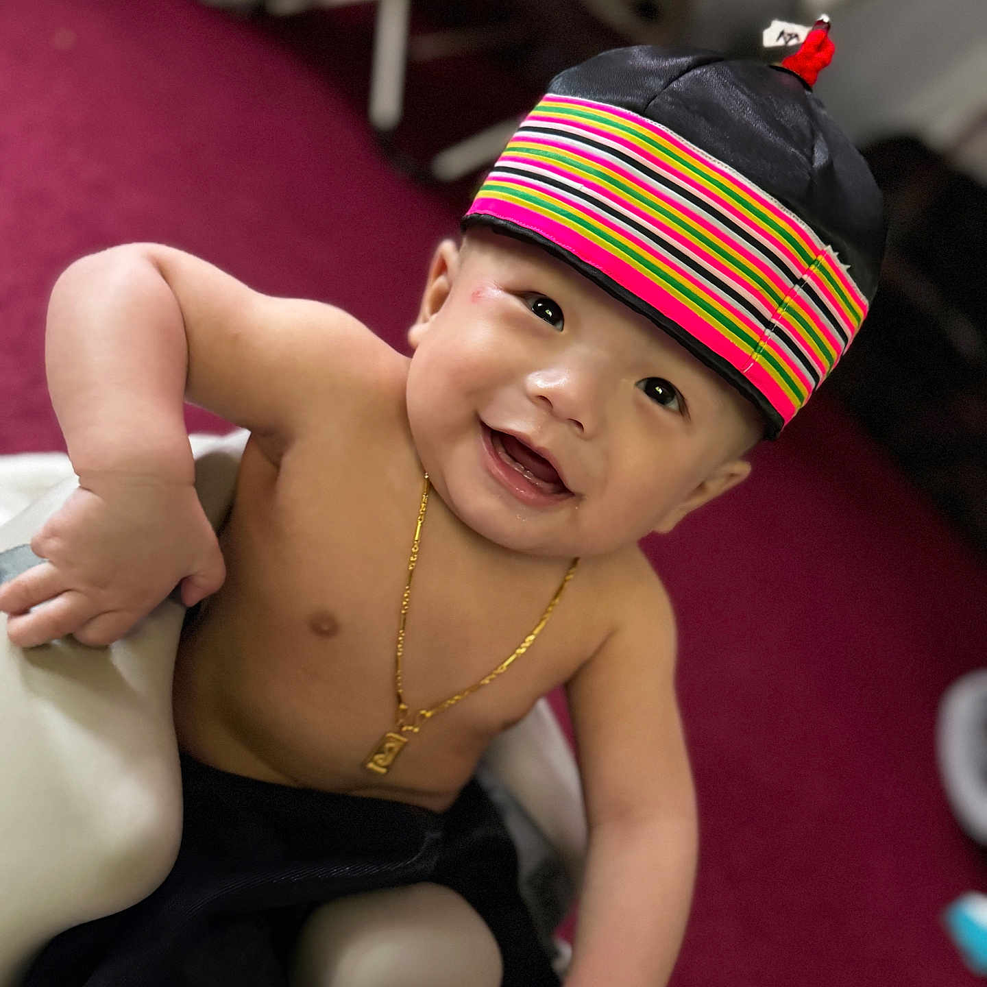 Luca joined the competition — help win amazing prizes! baby, child, smiling, hat, colorful_hat, jewelry, necklace, happy, indoors, carpet, chair, skin, portrait, cute, person, young_child, sitting, black_pants, expression, closeup