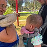 baby, child, person, woman, man, chicken, bird, animal, baby_carrier, outdoor, park, sunlight, glasses, fence, grass, trees, blue_shirt, casual_clothing, interaction, smile