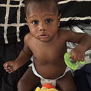Devon joined the competition — help win amazing prizes! baby, child, infant, portrait, sitting, bed, blanket, diaper, toy, plush_toy, green_toy, colorful, curious, serious_expression, indoors, bedroom, dark_skin, bare_chest, hands, feet