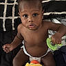 baby, child, infant, portrait, sitting, bed, blanket, diaper, toy, plush_toy, green_toy, colorful, curious, serious_expression, indoors, bedroom, dark_skin, bare_chest, hands, feet