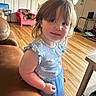 toddler, child, blue_dress, indoor, wooden_floor, couch, furniture, toys, pink_chair, door, shoes, person, young_child, curly_hair, casual_clothing, smiling, home_interior, flooring, small_chair, portrait
