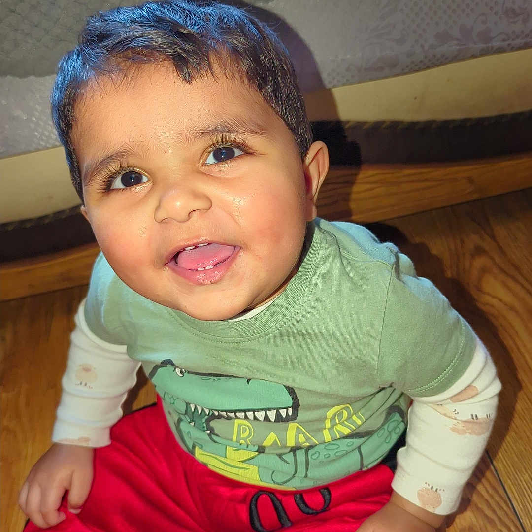 Umar joined the competition — help win amazing prizes! baby, bed, child, clothing, cute, dinosaur_print, face, floor, green_shirt, happy, home, indoor, long_sleeves, person, red_pants, sitting, smiling, toddler, white_socks, wooden_floor