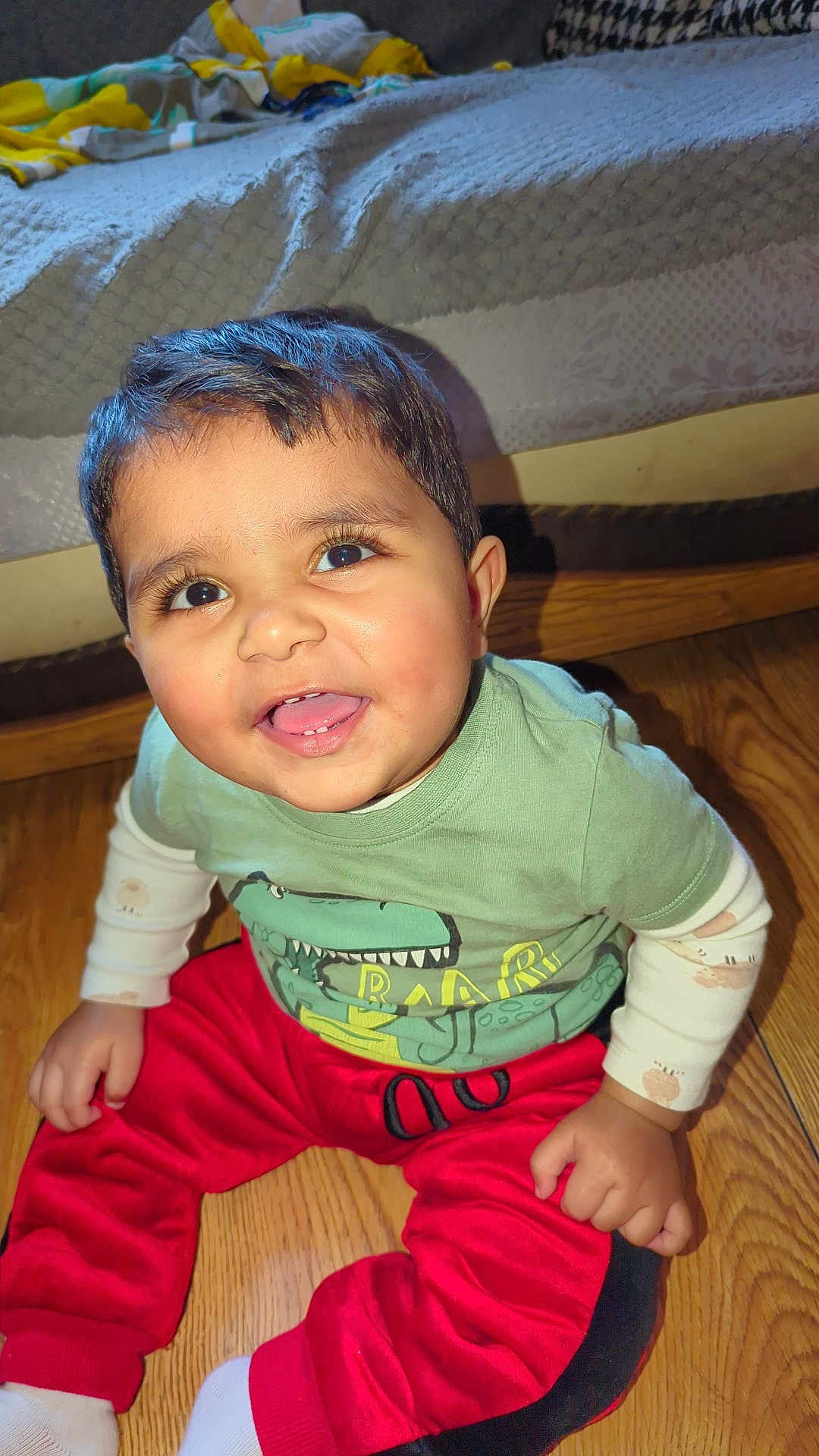 Umar joined the competition — help win amazing prizes! toddler, child, smiling, sitting, indoor, floor, wooden_floor, clothing, green_shirt, red_pants, dinosaur_print, cute, happy, face, person, baby, long_sleeves, white_socks, home, bed