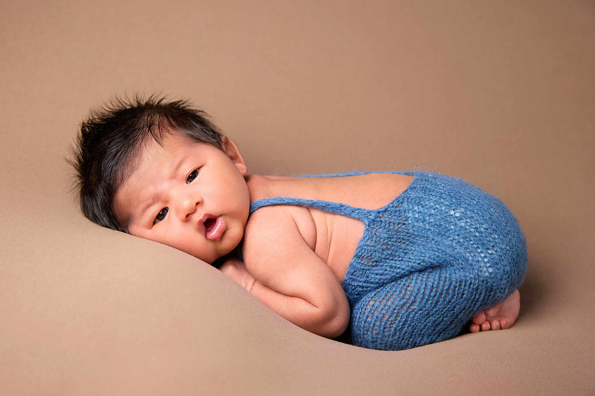 Jaycob is registered to the contest to win money with this photo: baby, infant, newborn, blue_outfit, knitted_clothing, soft_texture, curly_hair, lying_down, beige_background, cute, chubby_cheeks, portrait, child, skin, face, expression, resting, small_feet, indoors, cozy