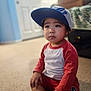 child, toddler, cap, baseball_cap, red_shirt, long_sleeves, sitting, carpet, bed, bedspread, suitcase, portrait, face, eyes, expression, indoors, soft_focus, bokeh, floor, hand