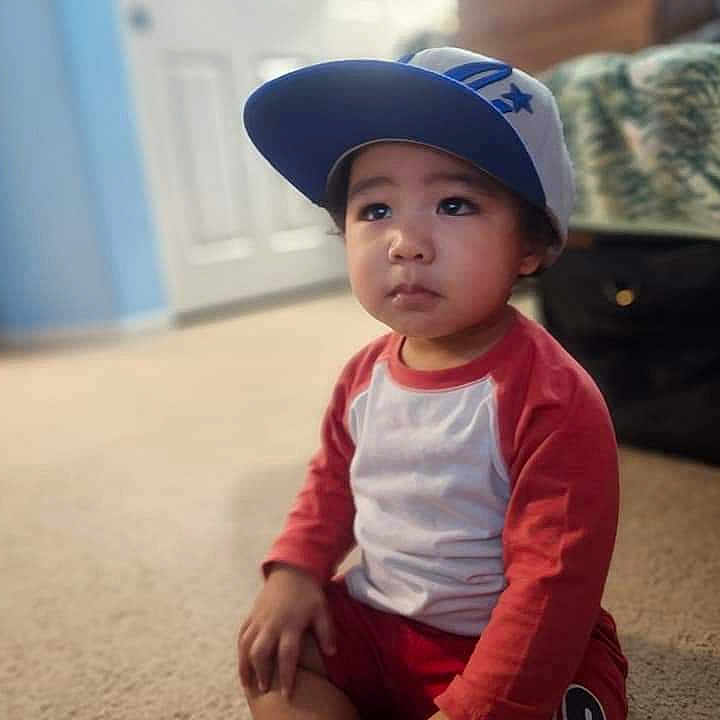 Johannes Clemente joined the competition — help win amazing prizes! baseball_cap, bed, bedspread, bokeh, cap, carpet, child, expression, eyes, face, floor, hand, indoors, long_sleeves, portrait, red_shirt, sitting, soft_focus, suitcase, toddler