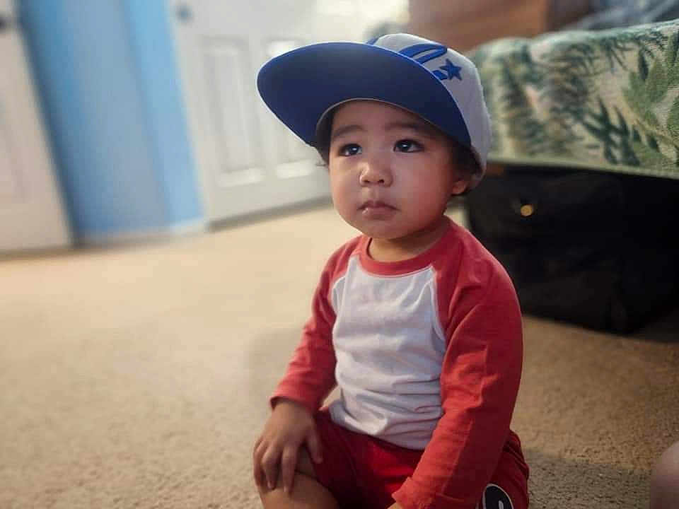 Johannes Clemente joined the competition — help win amazing prizes! child, toddler, cap, baseball_cap, red_shirt, long_sleeves, sitting, carpet, bed, bedspread, suitcase, portrait, face, eyes, expression, indoors, soft_focus, bokeh, floor, hand