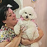 Tina a rejoint le concours — aidez-le/la à gagner de superbes lots ! woman, person, dog, white_dog, fluffy, curly_fur, pet, cuddling, smiling, indoors, portrait, short_sleeves, floral_shirt, pink_harness, dog_collar, thermostat, wall, home, seated, happy