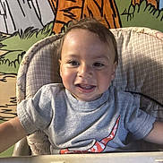 Mateo is registered to the contest to win money with this photo: toddler, child, high_chair, smiling, gray_tshirt, nike_logo, indoor, colorful_background, cartoon_mural, happy, face, short_hair, portrait, baby, seated, clothing, person, cute, playful, expression