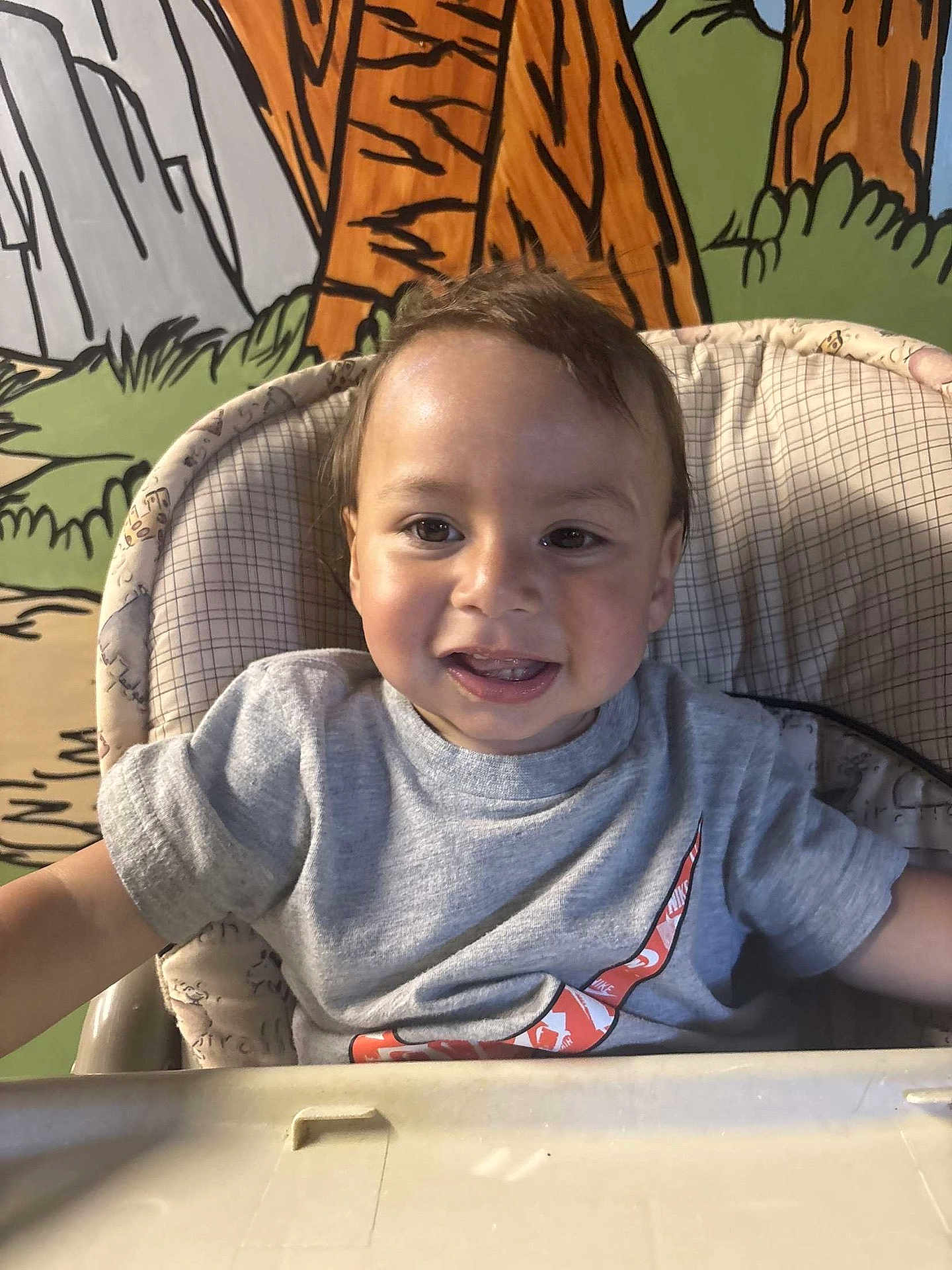 Mateo is registered to the contest to win money with this photo: toddler, child, high_chair, smiling, gray_tshirt, nike_logo, indoor, colorful_background, cartoon_mural, happy, face, short_hair, portrait, baby, seated, clothing, person, cute, playful, expression