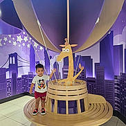 Langston is registered to the contest to win money with this photo: toddler, child, mickey_mouse, shorts, sneakers, wooden_structure, hot_air_balloon, giraffe_cartoon, purple_background, cityscape, stars, indoor, floor_tiles, bench, smiling, play_area, decorative, cute, fun, posing